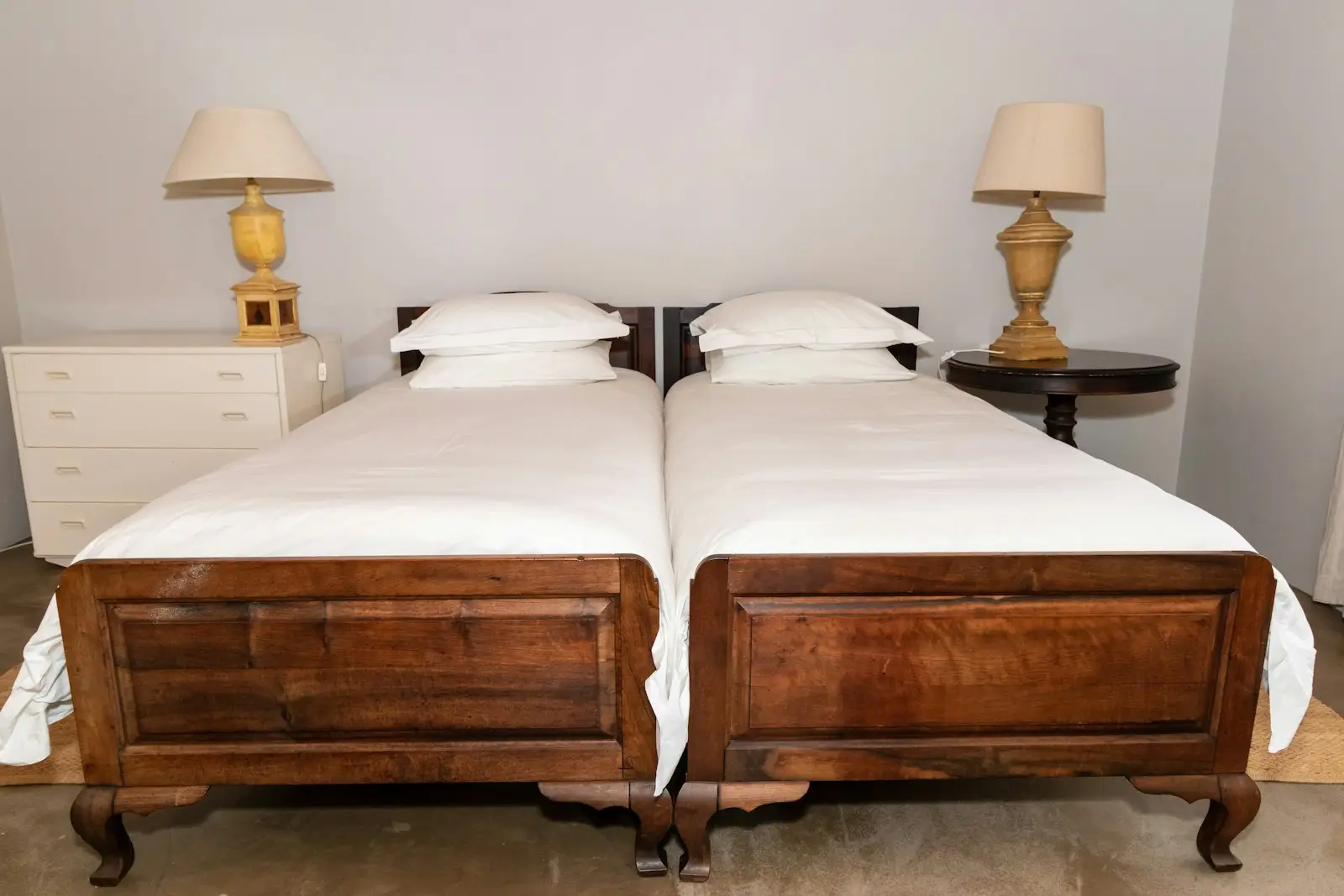 Dos camas de madera con piecera de paneles y patas torneadas, junto a veladores con lámparas